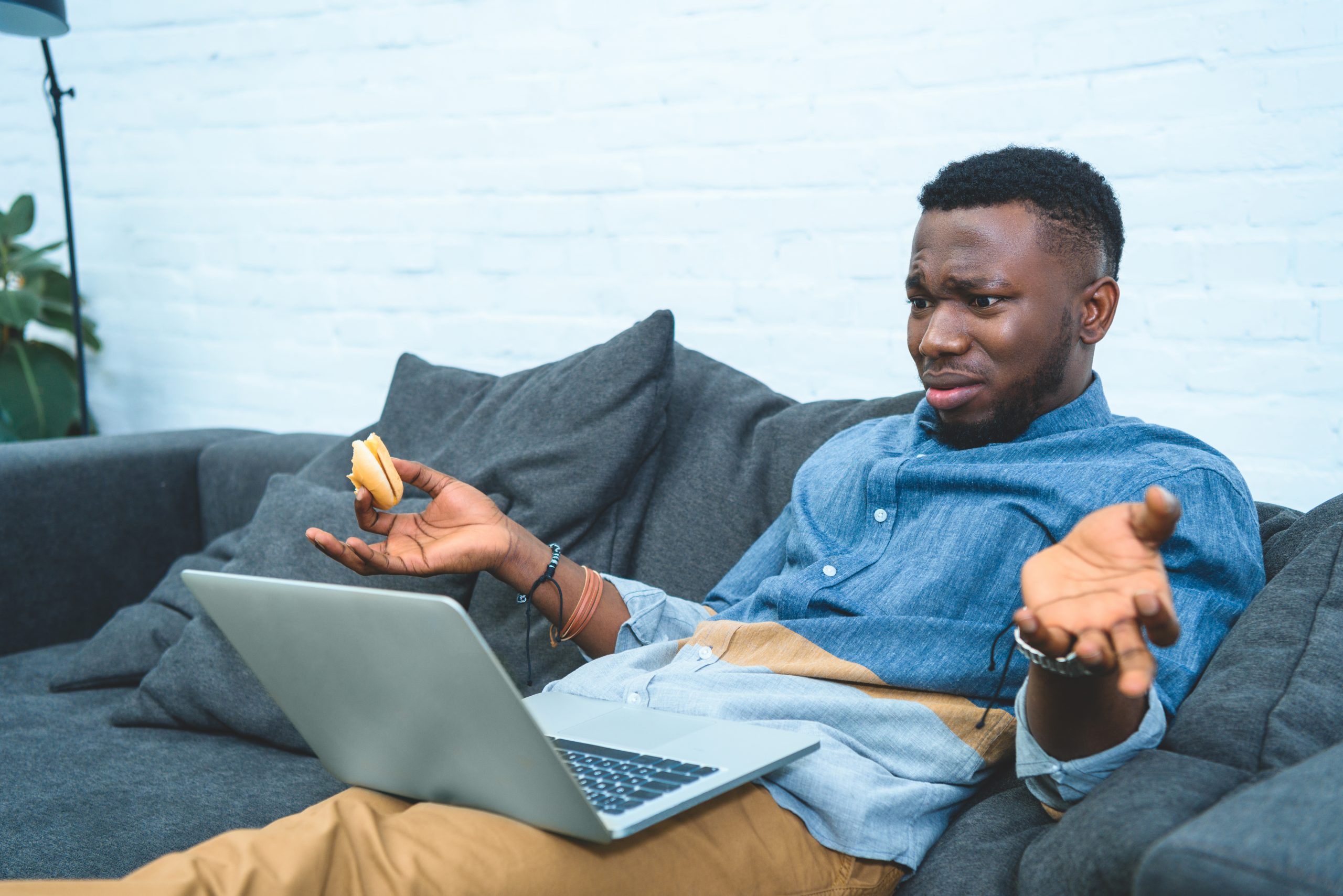 Confused african american man working on laptop while sitting on sofa and eating hamburger image of a confused consumer due to common marketing fails